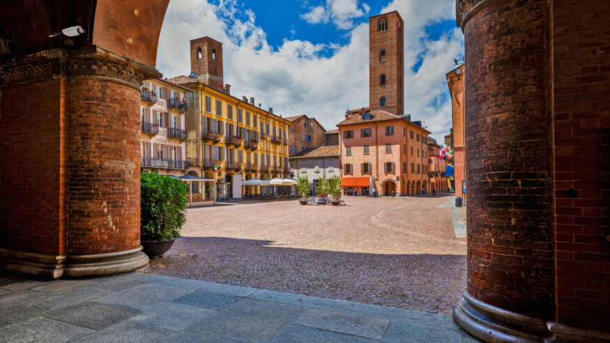 View of cobblestone town square among old houses and medieval towers under beautiful sky in Alba, Piedmont, Northern Italy.