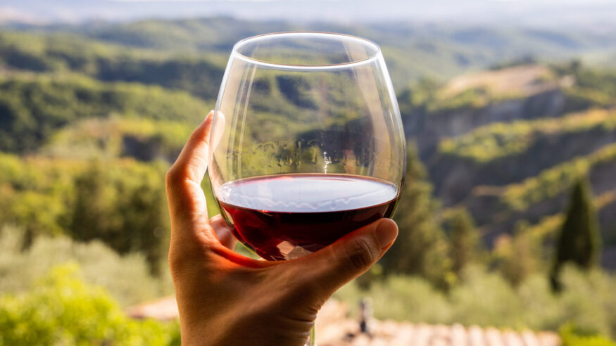 Woman holding a glass of red wine with beautiful landscape of Italy in a background on a sunny day. View from the window.